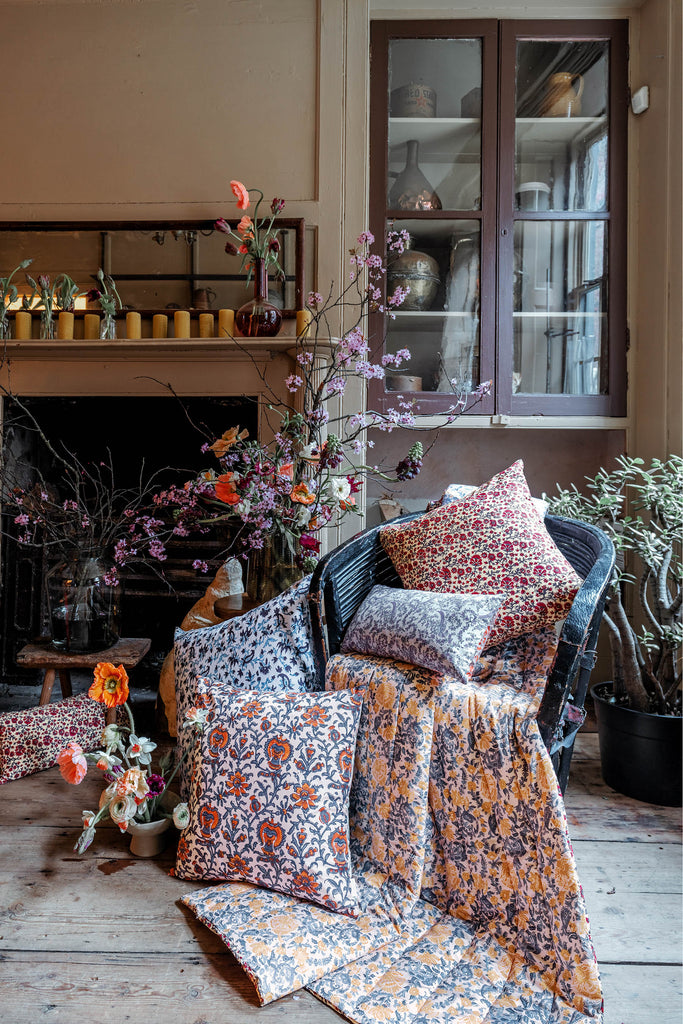 Living room with chairs decorated with red, blue, yellow, pink, green and blue cushions and quilts. Floral arrangements are laid out around the cushions on the wooden floors.