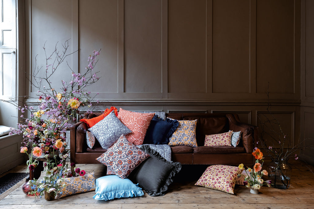 Green, yellow, blue, pink, red floral patterned silk cushions set on a brown leather sofa and on a wooden floor with a floral arrangement around the sofa