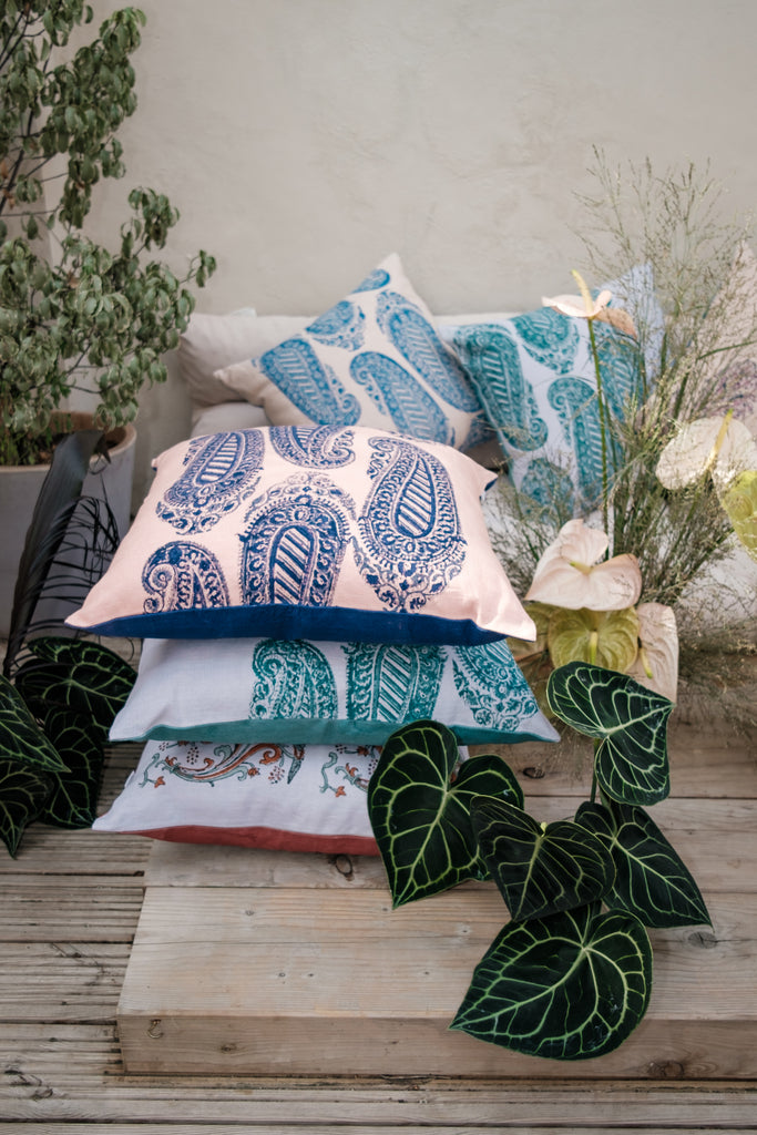 Garden with outside seating with colourful block printed linen cushions on it, coloured beige, green, blue, purple and pink surrounded by flowers and trees.