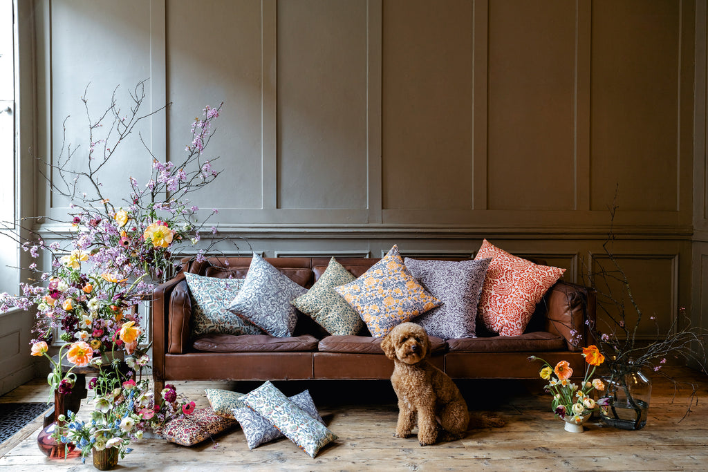 Green, yellow, blue, pink, red floral patterned silk cushions set on a brown leather sofa and on a wooden floor with a floral arrangement around the sofa and a brown poodle dog sitting on the wood floor. 