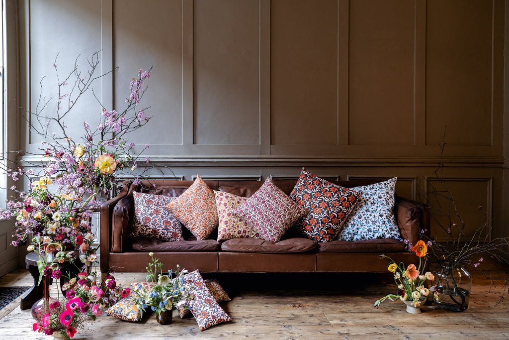 Green, yellow, blue, pink, red floral patterned silk cushions set on a brown leather sofa and on a wooden floor with floral arrangements around the sofa