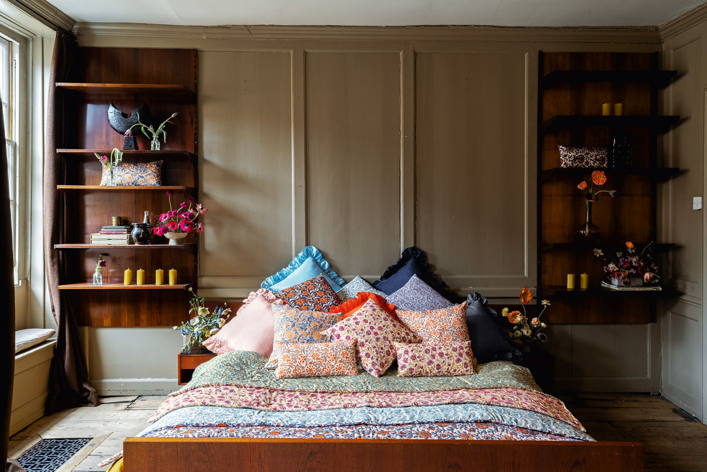 Green, yellow, blue, pink, red floral patterned silk cushions and quilts set on a bed in a bedroom with floral arrangements sitting on the bedside tables and shelves. 