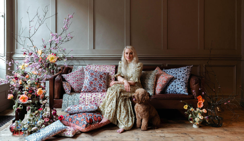 felicity founder of daughters of gaea wearing long yellow patterned dress sitting on brown leather sofa surrounded by floral cushions and flower arrangements. brown poodle dog sitting on the floor. 