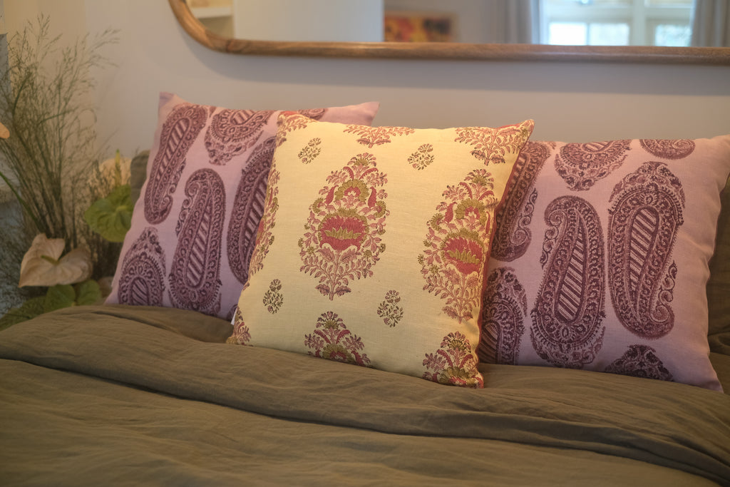 Three block printed linen cushions on a bed with colours of yellow, pink and purple with plants on the bedside table.  