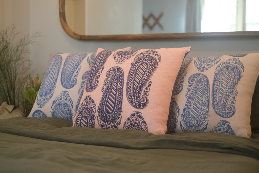 Three block printed linen cushions coloured pink, beige and blue on a bed with linen green sheets with flowers on the bedside table.