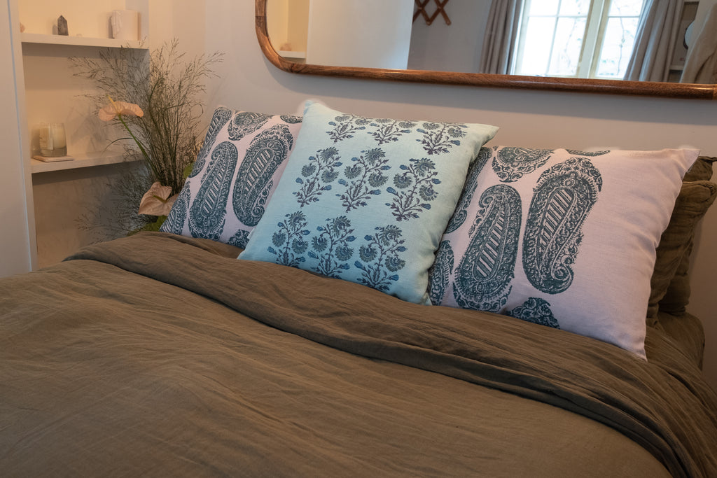 Purple, blue and green block printed linen cushions sit on a bed with flowers on the bedside table and a mirror above. 