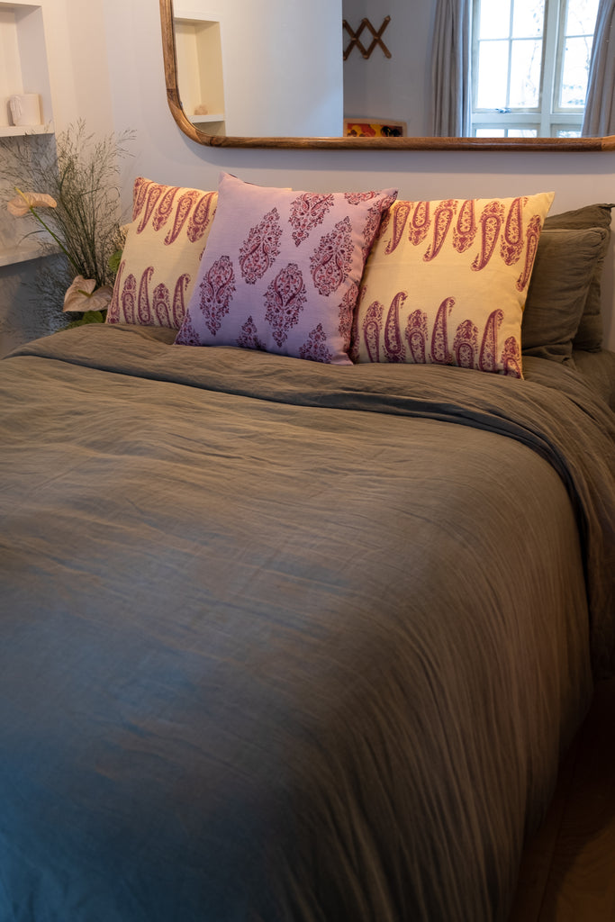 Three block printed linen cushions coloured pink, purple and yellow on a bed with linen green sheets with flowers on the bedside table.