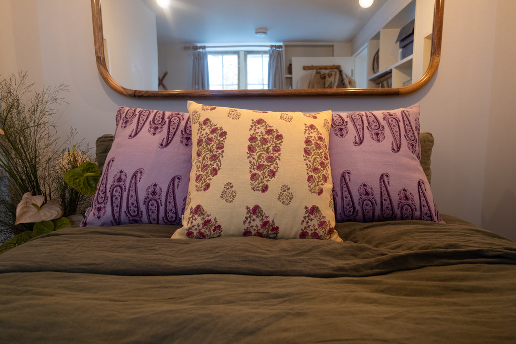 Three block printed linen cushions on a bed with purple, pink and yellow colours. Flowers by the bed and mirror above it. 